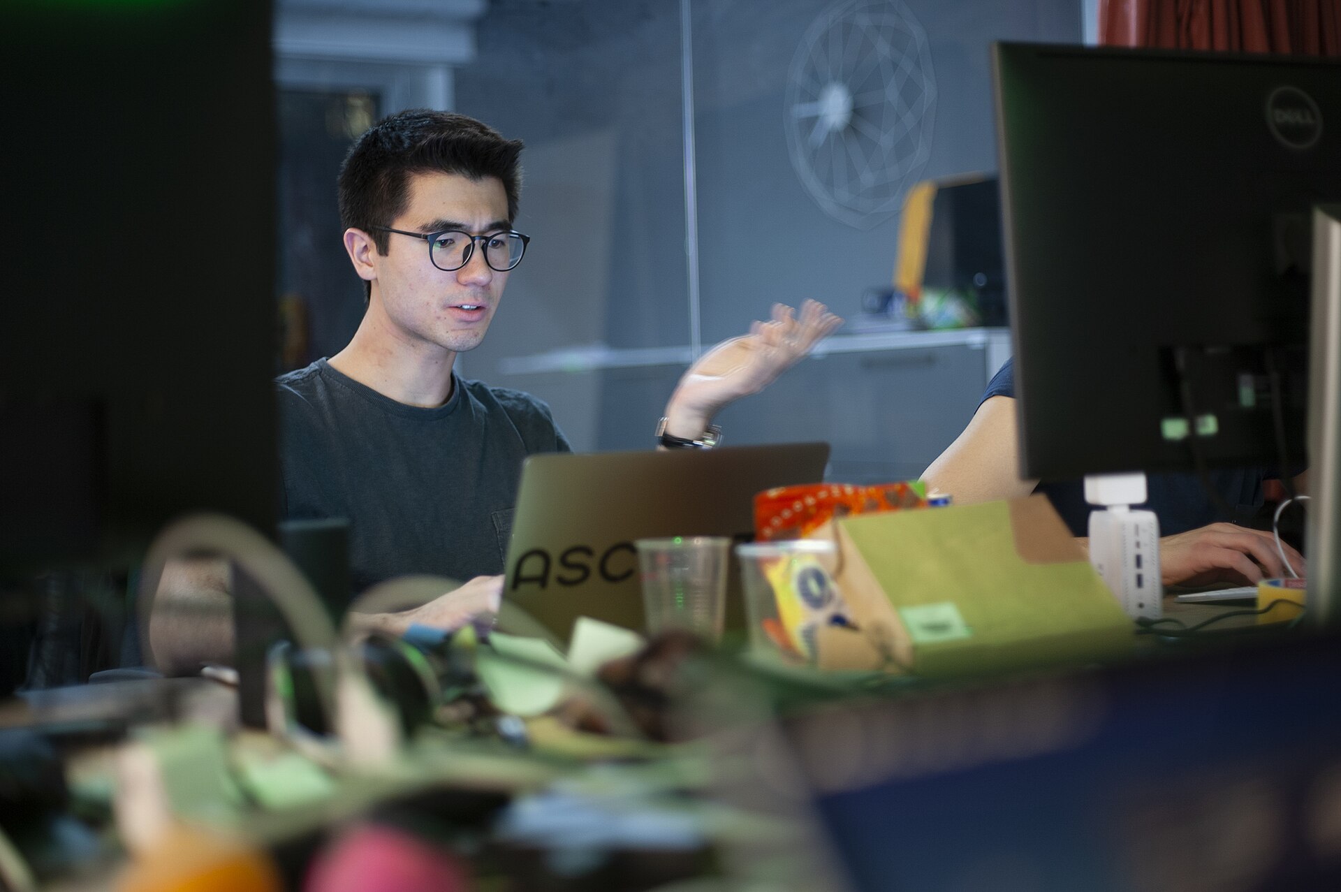 A student in glasses talking across a laptop at a hackathon, monitors either side